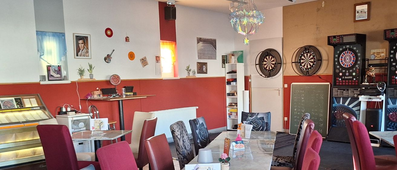 A dining area with a marble table and chairs, a projector, and dartboards. The room has a red wall with a picture frame, a plant, and a shelf with items.