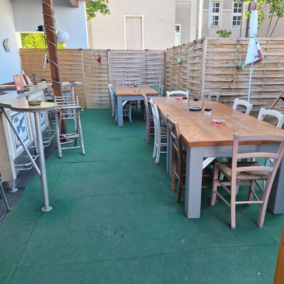 Ein Outdoor-Terrasse mit mehreren Holztischen und Stühlen, einem grün gefliesten Boden und Holzzäunen. Eine Lampe und ein zusammengeklappter Regenschirm sind ebenfalls vorhanden.