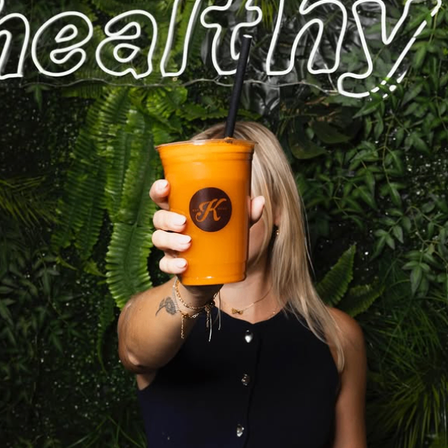 Eine Frau in einem schwarzen Oberteil hält einen Becher mit orangefarbenem Saft und einem schwarzen Strohhalm. Das Wort 'gesund' ist in Weiß über ihr geschrieben.