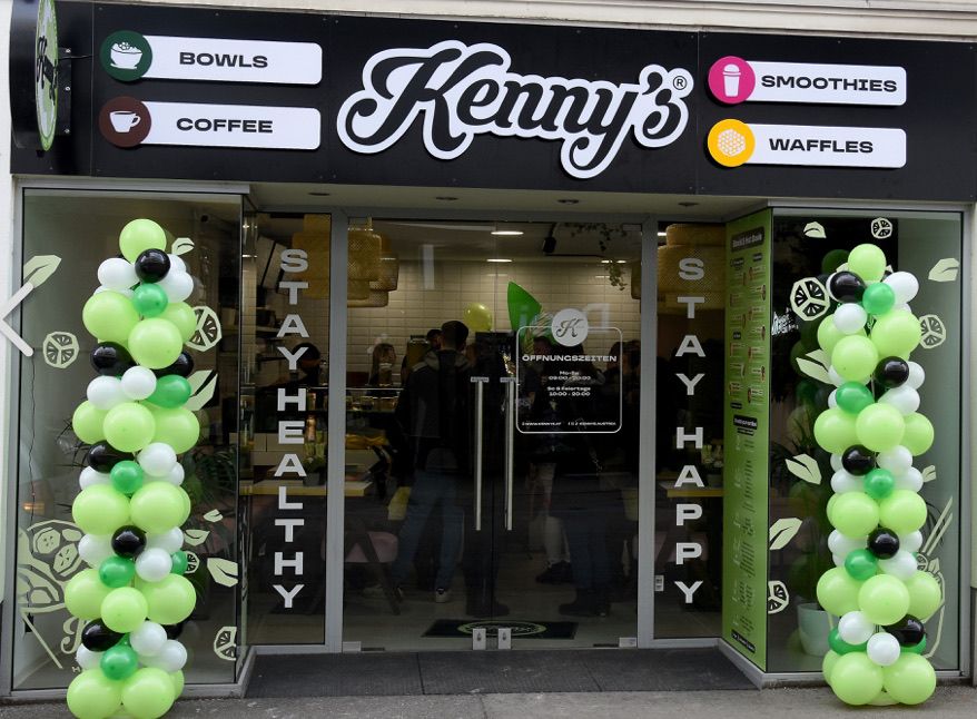 Vorne die Kenny's-Filiale mit grünen und weißen Luftballons auf beiden Seiten des Eingangs. Der Laden verkauft Schalen, Kaffee, Smoothies und Waffeln.