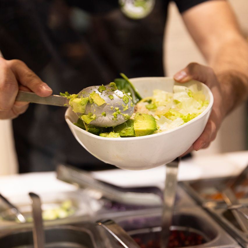 Eine Person in einem schwarzen T-Shirt fügt einer Salatschüssel Avocado mit einem Löffel hinzu. Die Schüssel enthält Salatgrün und es sind zusätzliche Lebensmittelvorbereitungsbehälter in der Nähe.