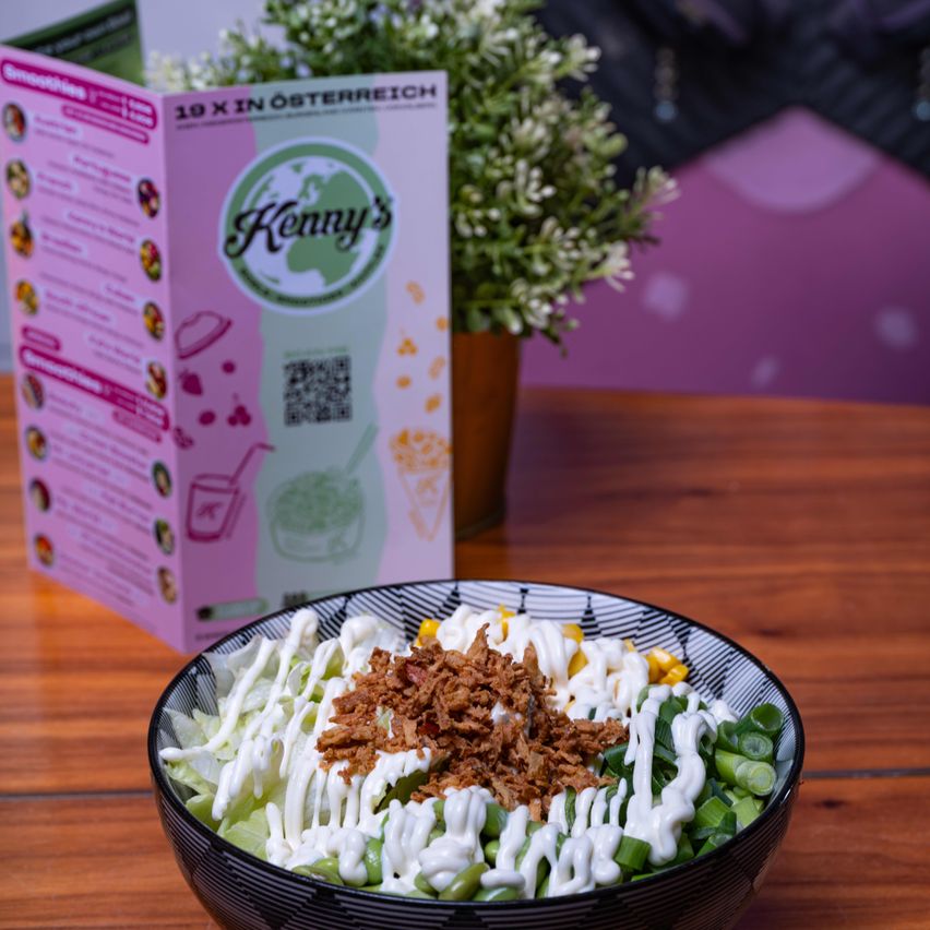 Ein Salatschüssel mit Speckstückchen, Mais und Dressing auf einem Holztisch. Dahinter befindet sich ein Menü für Kenny's, mit einer Topfpflanze in der Nähe.