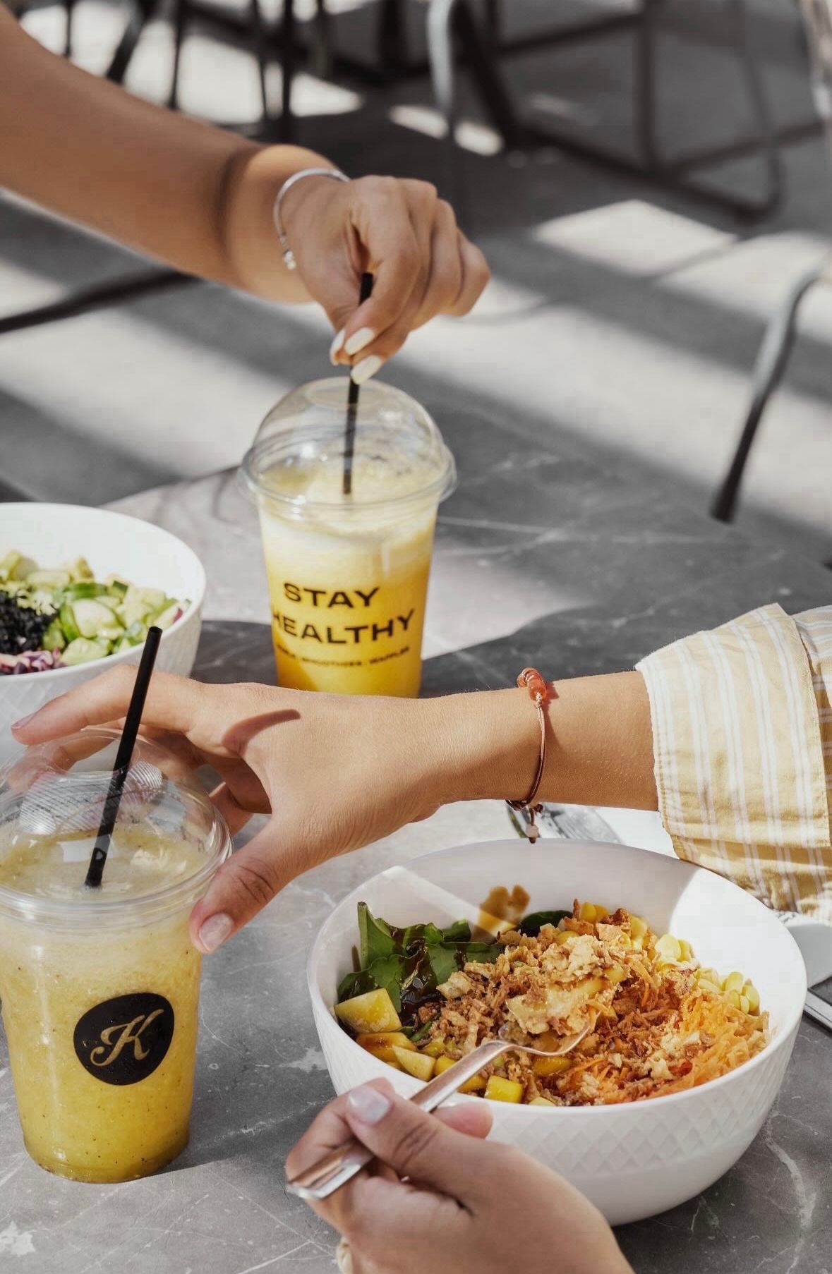 Zwei Hände greifen nach Schüsseln mit Essen und Getränken auf einem Tisch. Eine Schüssel enthält Salat, und die anderen beiden enthalten ein gelbes Getränk mit einem schwarzen Strohhalm. Ein Armreif befindet sich am Handgelenk.