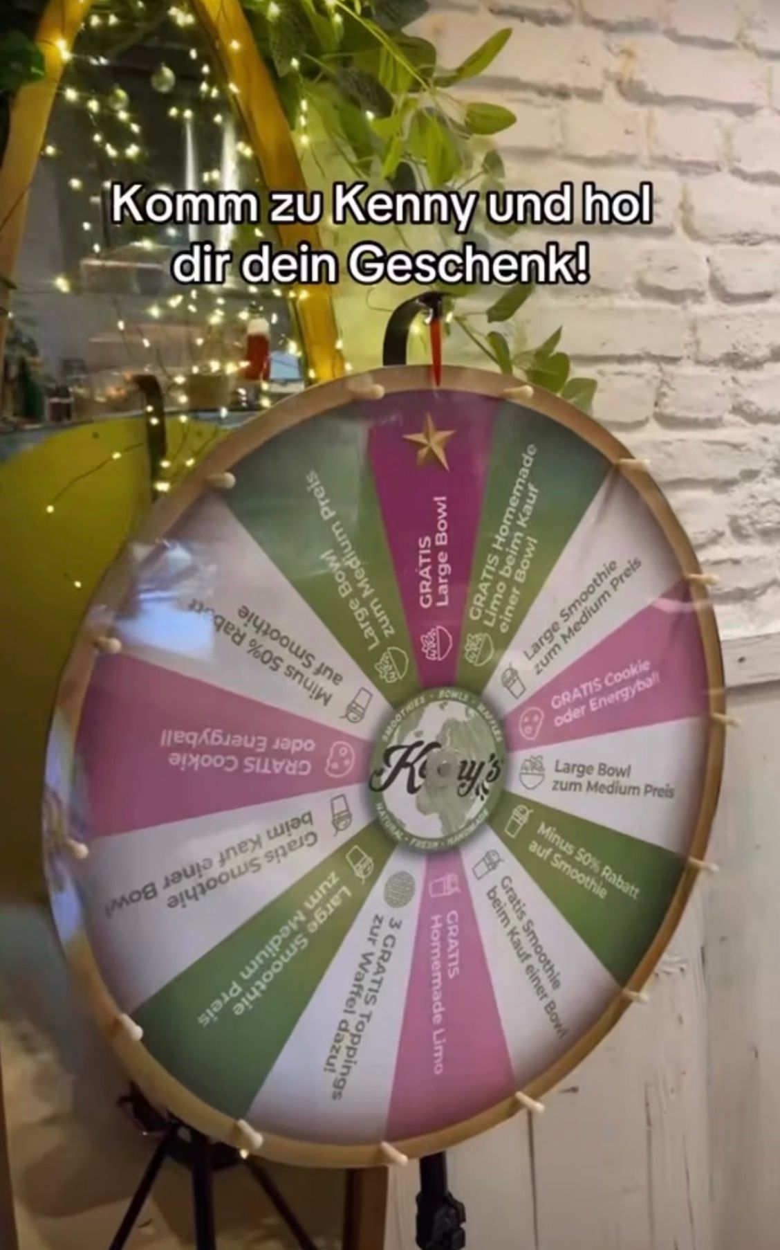 A colorful spinning wheel offers various prizes, including a large smoothie bowl, a medium smoothie, a gratis cookie, and a gratis energyball. The wheel is displayed against a white brick wall.
