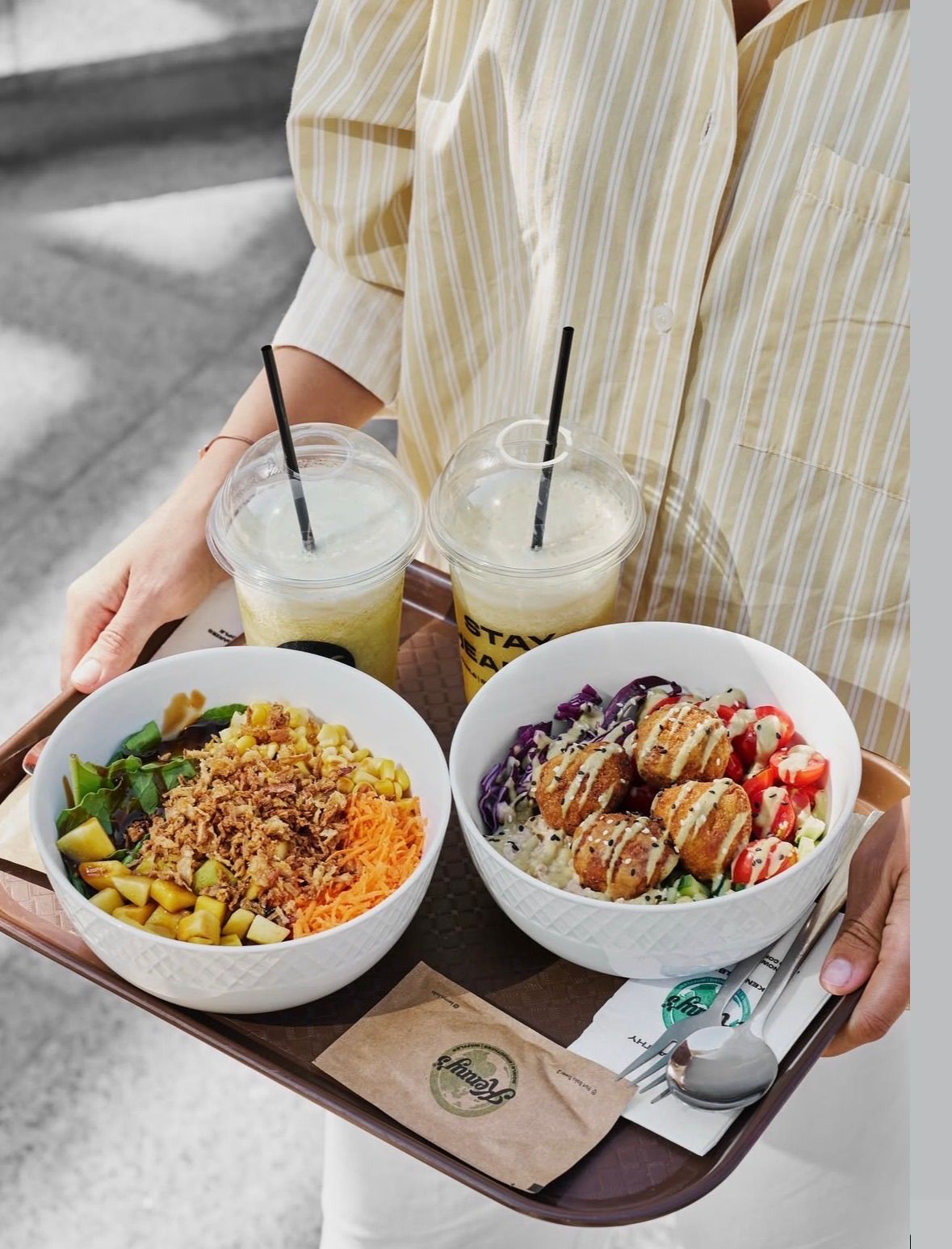 Eine Person hält einen Tablett mit zwei Salatschüsseln und zwei Getränken, eines mit einem Strohhalm.