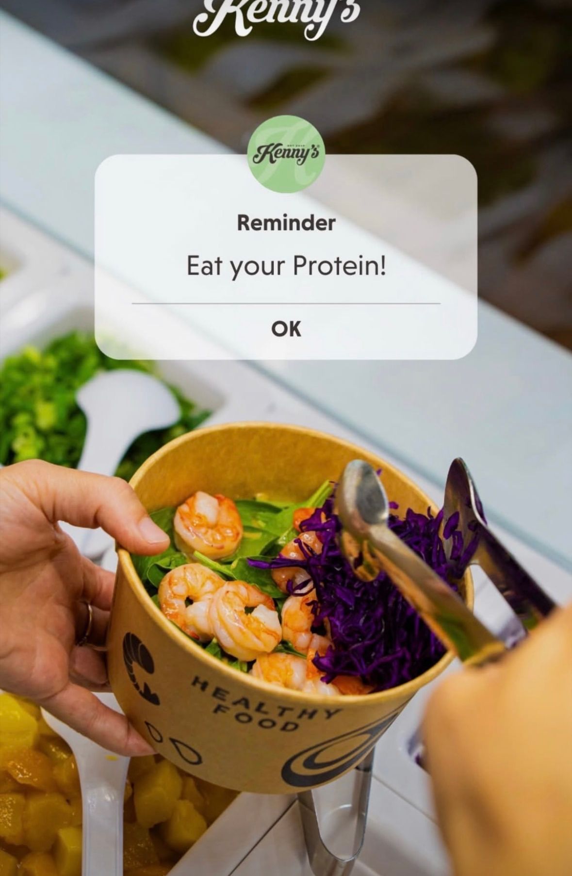 Eine Hand hält eine Schüssel mit Garnelensalat und roter Kohl. Eine Erinnerungsnachricht lautet 'Iss dein Protein!' mit einer OK-Schaltfläche.