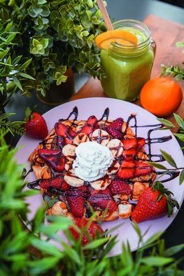 Ein Teller mit Waffeln, Schlagsahne, Erdbeeren, Bananen und Schokoladensauce steht neben einem Glas grünem Smoothie, einer Orange und Pflanzen.