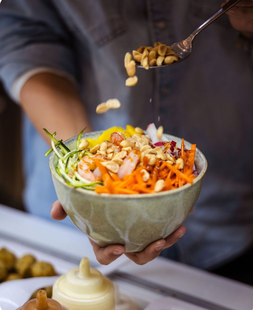 A person is holding a bowl of salad with chopped vegetables, shrimp, and peanuts. They are sprinkling peanuts on top of the salad.