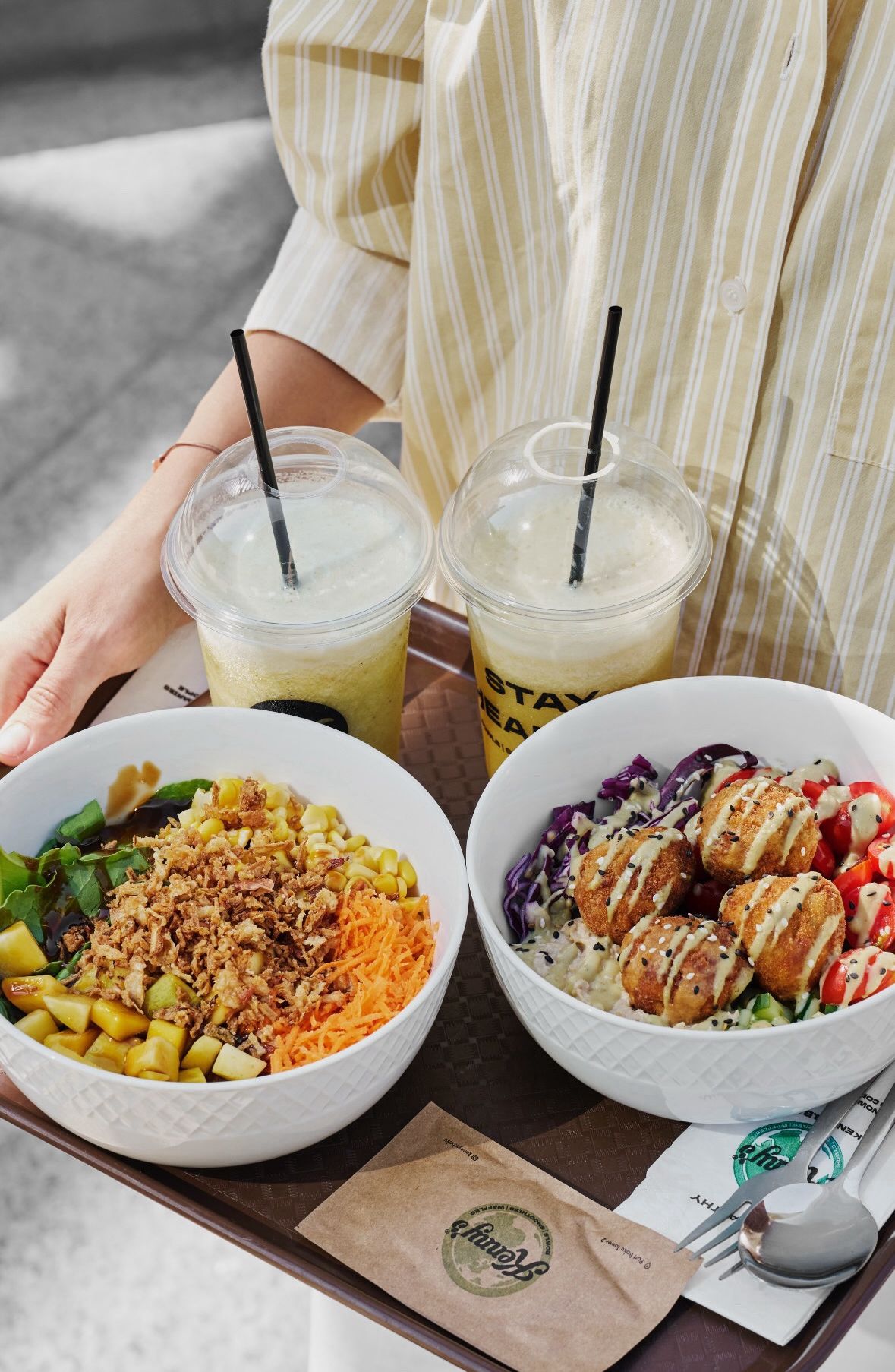 Two bowls of food and two cups of drinks on a tray. One bowl contains salad with veggies, fruits, and fried balls, while the other has vegetables, fruits, and fried balls. The cups have straws.