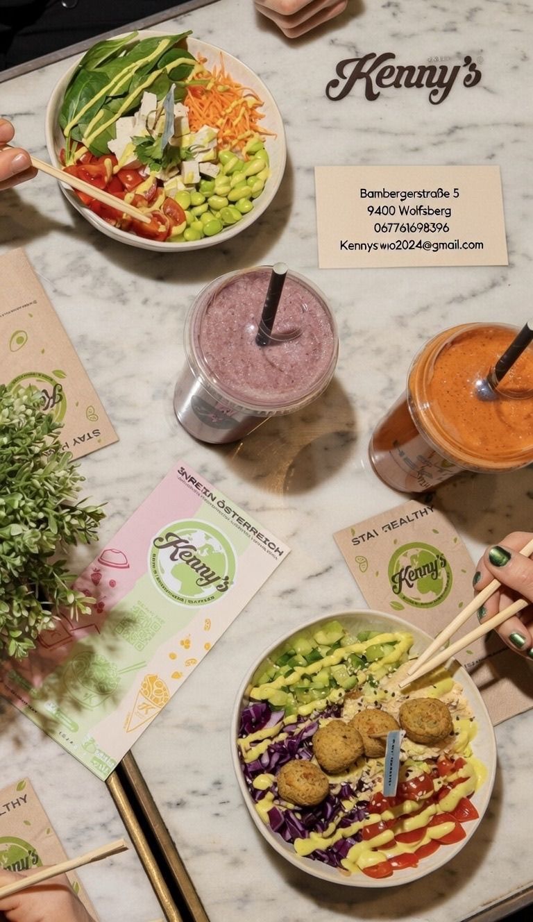 A table with various healthy food and drinks, including a bowl of salad, two smoothies, and a plant. There is also a menu card and a business card with the name Kenny's.