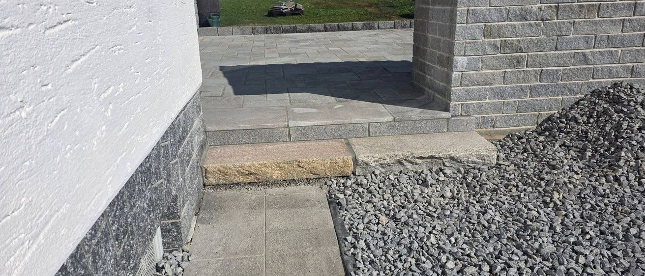 Ein gepflasterter Weg mit Steinstufen führt zu einem Rasenbereich. Schatten werden auf den Boden geworfen. Kies und Steine umgeben den Weg.