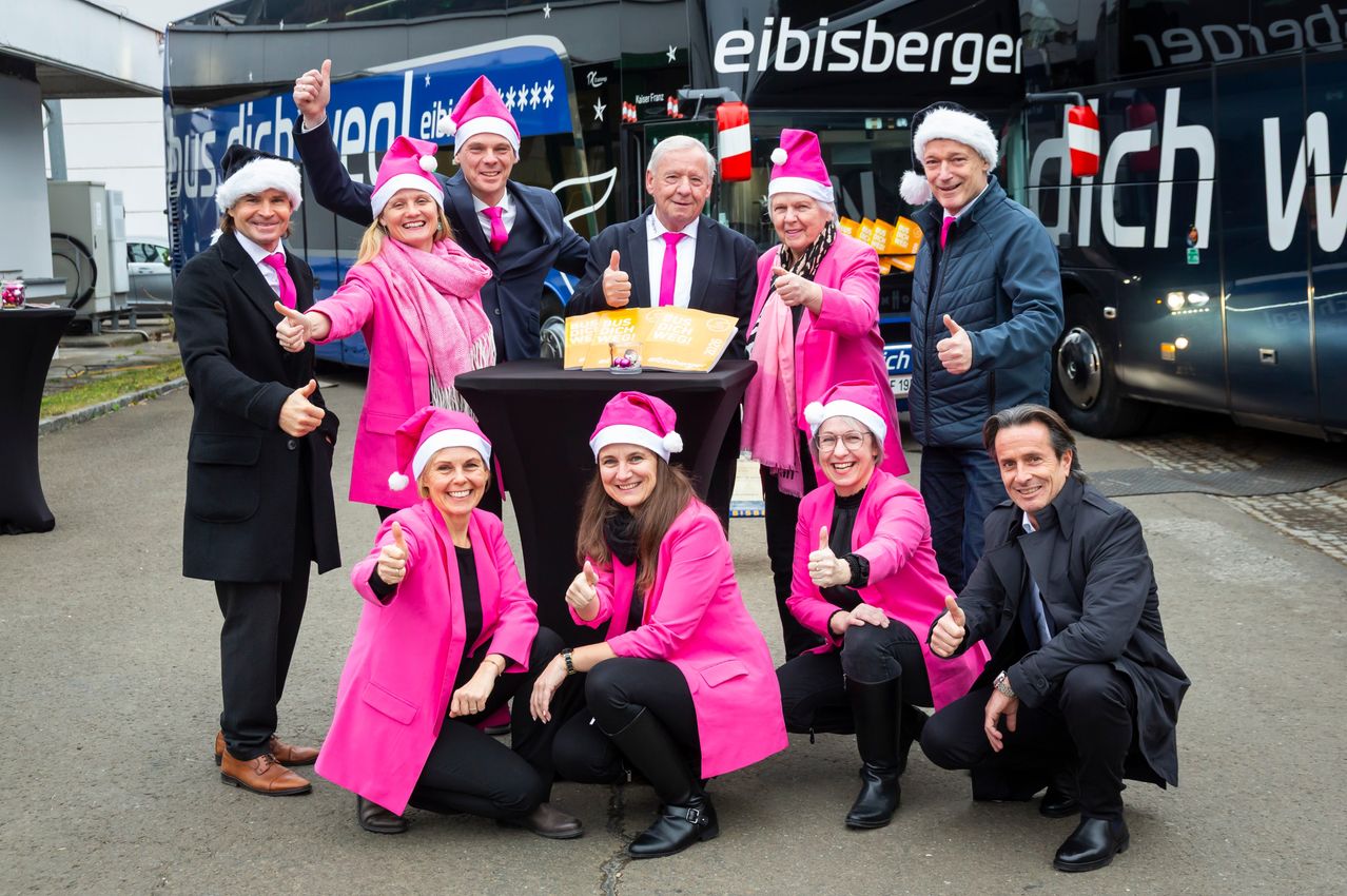 Eine Gruppe von Menschen in rosa Mänteln und Mützen posiert für ein Foto vor einem Bus mit dem Wort eibsiberger. Sie alle lächeln und geben Daumen hoch.