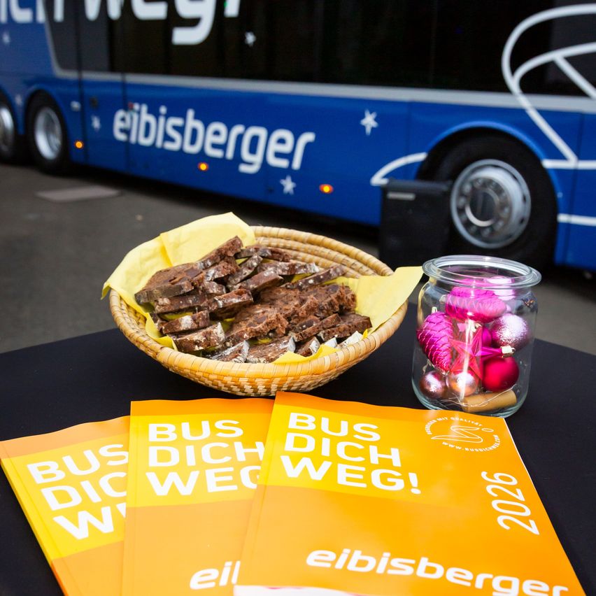 Auf einem Tisch liegt ein orangefarbenes Pamphlet mit dem Text 'BUS DICH WEG!' und '2020'. Ein Korb mit Brot und ein Glas mit Süßigkeiten steht vor einem blauen Bus.
