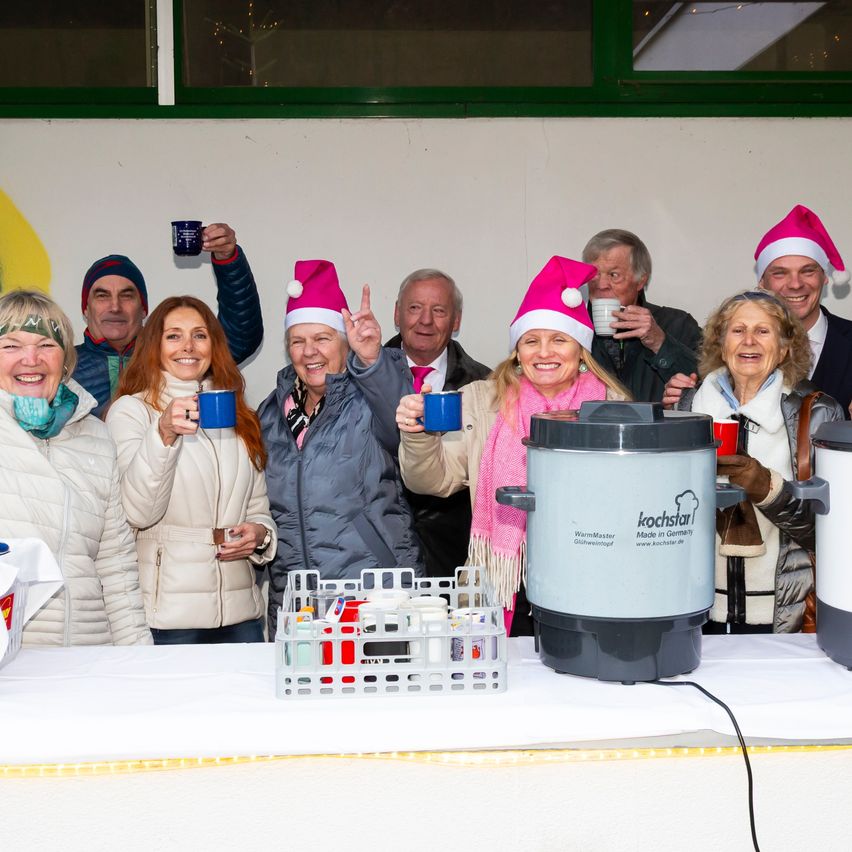 Eine Gruppe von Menschen in Weihnachtsmützen und Winterkleidung hält blaue Tassen und steht hinter einem Tisch mit einem Kaffeespender in einer festlichen Umgebung.