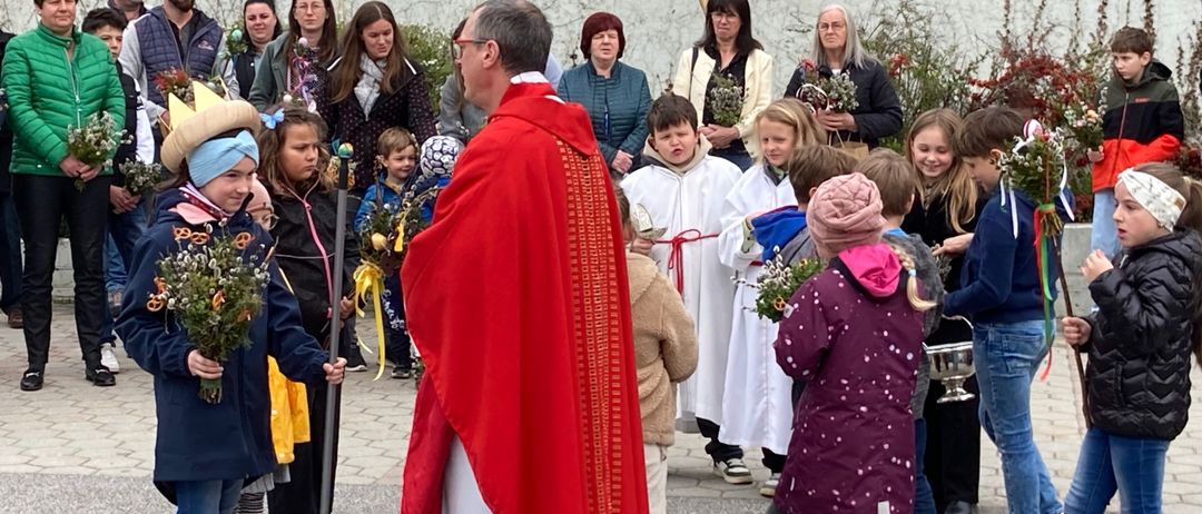 Ein Priester in rotem Gewand und mit Brille spricht zu einer Gruppe von Kindern und Erwachsenen, die Blumen halten, in einer Außenumgebung.