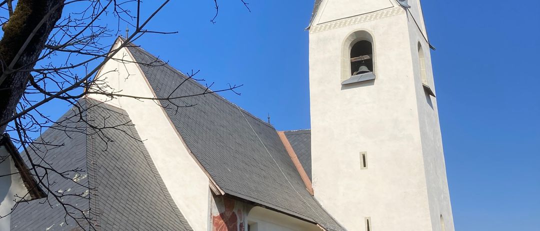 Eine Kirche mit einem hohen Turm und grauem Dach, geschmückt mit einem roten Wandbild, steht unter einem klaren blauen Himmel.