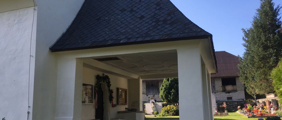 Das Äußere einer Kirche mit einem schwarzen Ziegeldach und weißen Wänden. Es hat eine Veranda mit Säulen, und im Hintergrund sind Pflanzen und Bäume sichtbar.