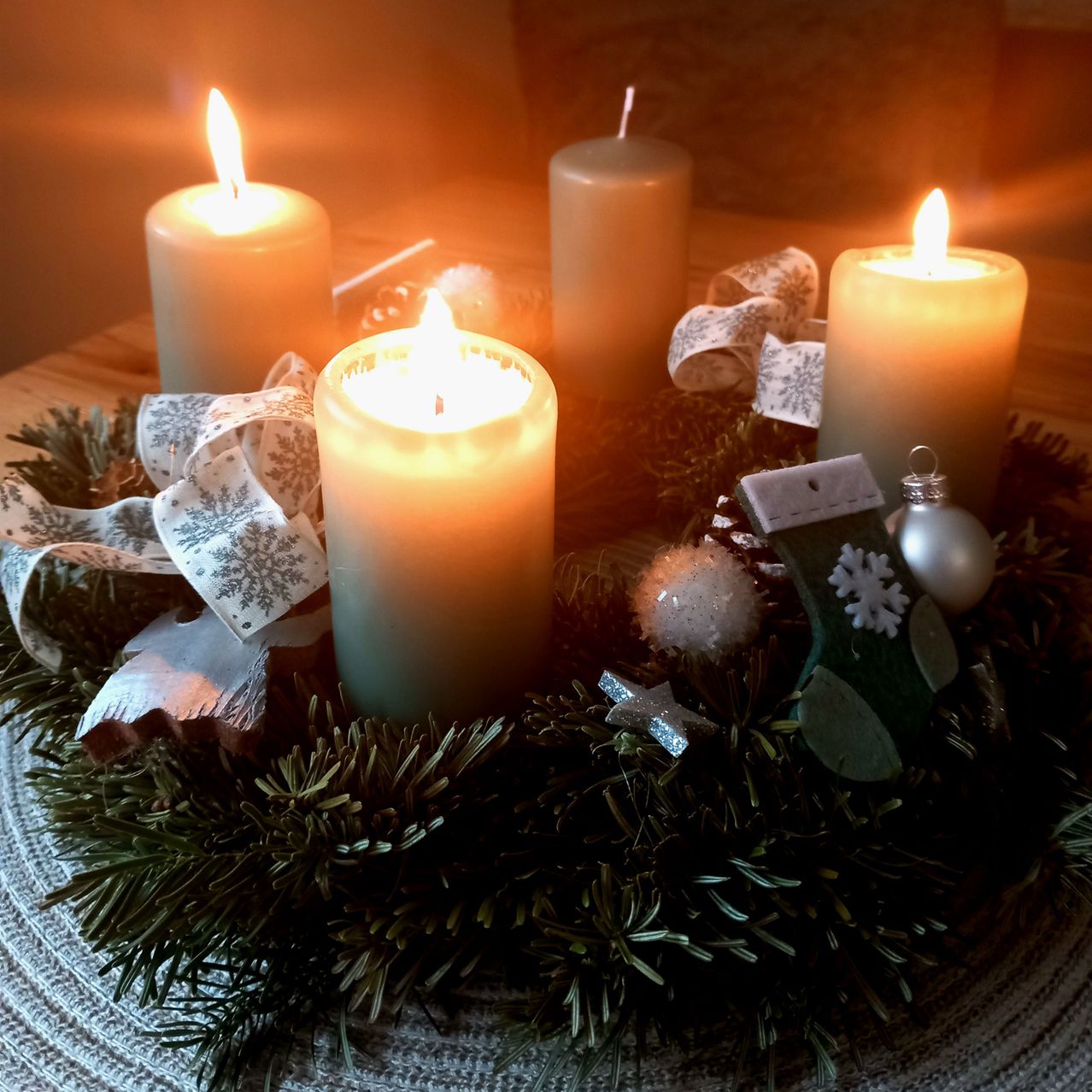 Ein Weihnachtskranz mit vier brennenden Kerzen, umgeben von Tannennadeln, Bändern und Ornamenten auf einem Tisch.