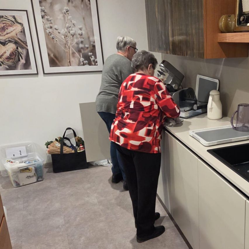 Zwei ältere Frauen arbeiten in einer Küche, eine steht vor einer Arbeitsplatte mit Küchengeräten. Ein Mann steht hinter ihr. In der Nähe befinden sich gerahmte Bilder an der Wand, ein Regal und verschiedene Gegenstände auf dem Boden.