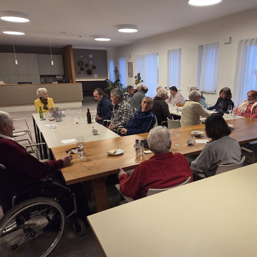 Eine Gruppe älterer Menschen sitzt um einen langen Tisch in einem Esszimmer, einige in Rollstühlen. Flaschen, Gläser und Teller sind auf dem Tisch. Eine Pflanze steht in der Ecke.
