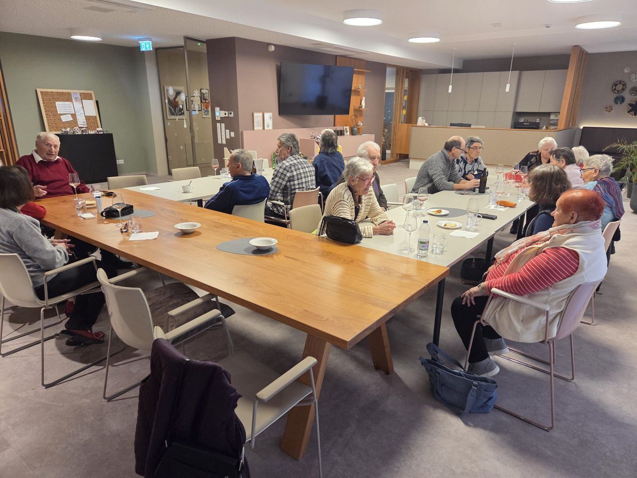 Eine Gruppe älterer Menschen sitzt um einen langen Tisch in einem Essbereich herum, möglicherweise bei einem Treffen. Sie trinken Wein und essen.
