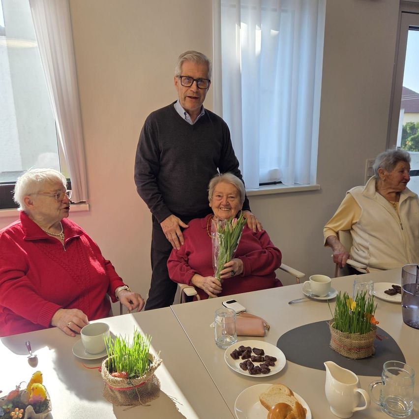 Drei ältere Menschen sitzen um einen Tisch. Ein Mann steht hinter einer Frau mit Blumen. Auf dem Tisch stehen Brot, Schokolade und Blumen. Der Raum hat weiße Wände, Fenster und Vorhänge.