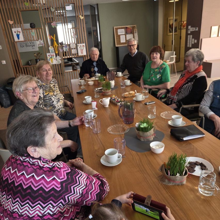 Mehrere ältere Menschen sitzen um einen Tisch herum, möglicherweise in einer Besprechung, mit Tassen, Tellern und Dekorationen. Eine Person hält ein Tablet.