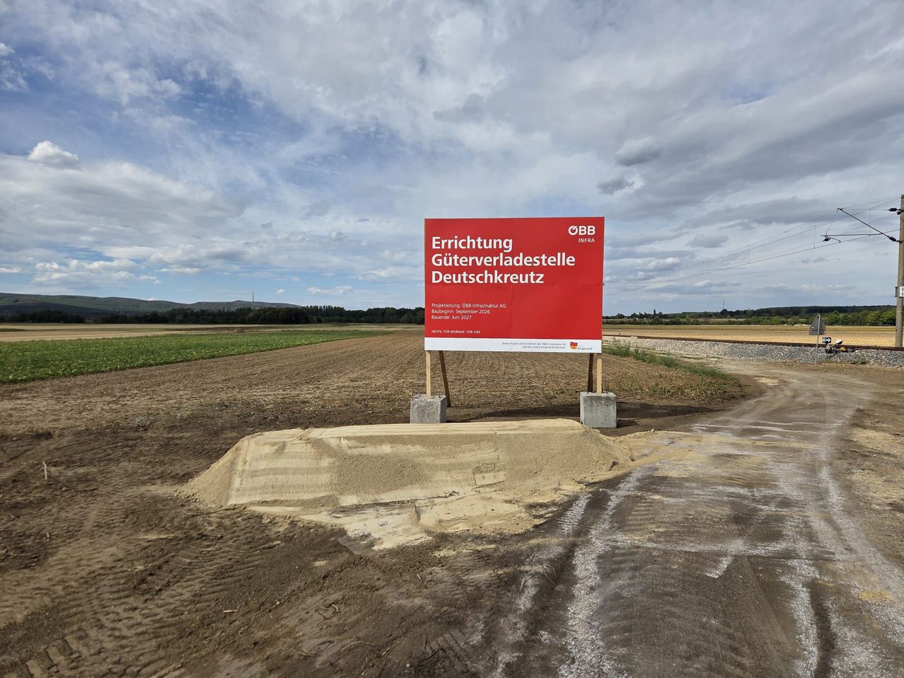 Ein rotes Schild auf einem Feld mit der Aufschrift 'Errichtung Güterverladestelle Deutschkreuz' und einem bewölkten Himmel im Hintergrund.