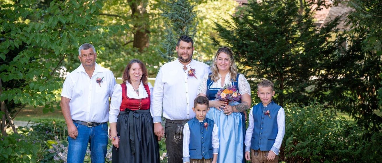 Eine Familie in traditioneller deutscher Kleidung posiert für ein Foto im Garten, mit zwei Männern, zwei Frauen und zwei Kindern, die zusammenstehen.