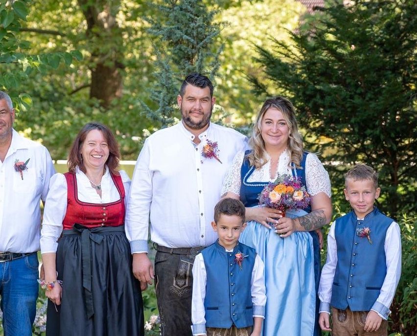 Eine Familie in traditioneller deutscher Kleidung posiert für ein Foto im Garten, mit zwei Männern, zwei Frauen und zwei Kindern, die zusammenstehen.