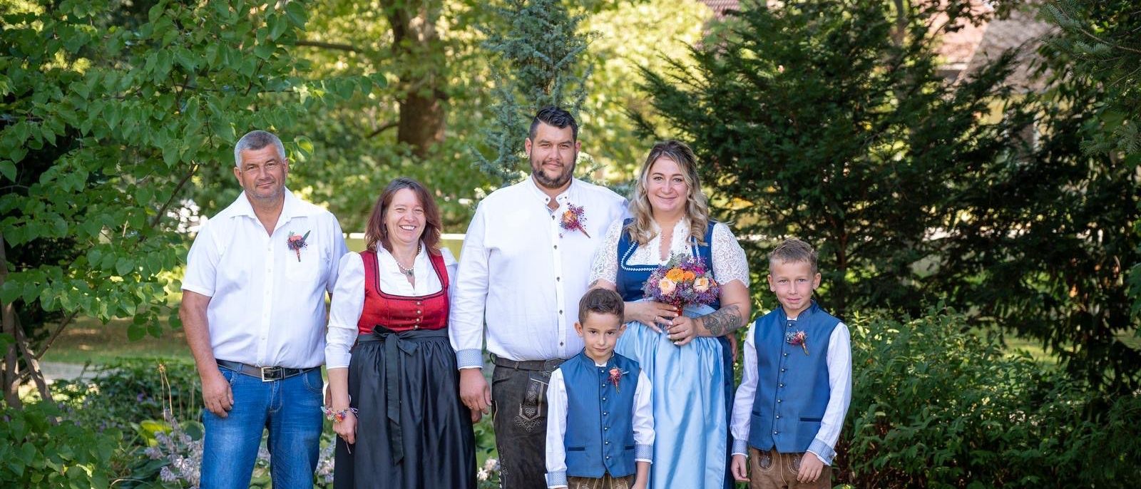 Eine Familie in traditioneller deutscher Kleidung posiert für ein Foto im Garten, mit zwei Männern, zwei Frauen und zwei Kindern, die zusammenstehen.