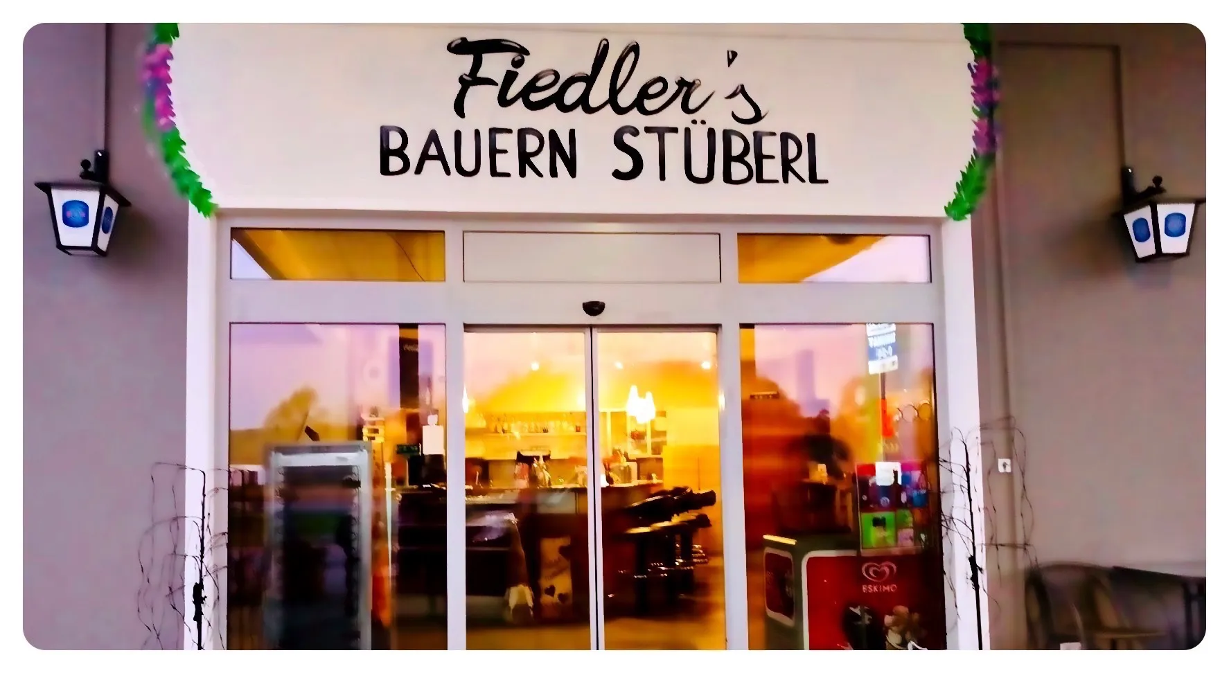 Die Fassade von Fiedler's Bayerischem Stuberl zeigt eine Glastür und Fenster, mit einem Schild darüber, das Fiedler's BAUREN STUBERL liest. Im Inneren ist eine Theke mit Stühlen und Regalen zu sehen.
