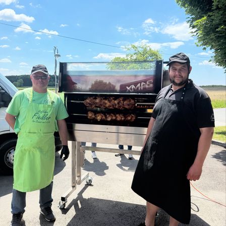 Zwei Männer in Schürzen stehen neben einem großen Grill im Freien. Ein Mann trägt eine grüne Schürze, der andere eine schwarze. Der Grill hat gekochtes Fleisch drin und ist mit 'XMAS' beschriftet. Ein weißer Lieferwagen und Bäume sind im Hintergrund.