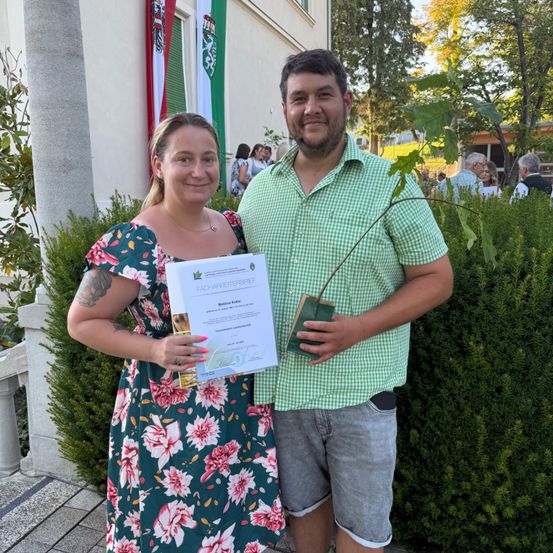 Ein Paar posiert für ein Foto draußen mit einem Zertifikat und einer kleinen Pflanze. Die Frau trägt ein Blumenkleid und der Mann ein grünes Hemd. Hinter ihnen sind Grünpflanzen und ein Gebäude mit Fahnen sichtbar.