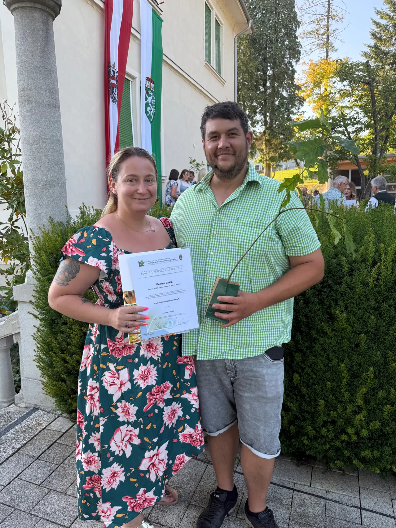 Ein Paar posiert für ein Foto draußen mit einem Zertifikat und einer kleinen Pflanze. Die Frau trägt ein Blumenkleid und der Mann ein grünes Hemd. Hinter ihnen sind Grünpflanzen und ein Gebäude mit Fahnen sichtbar.