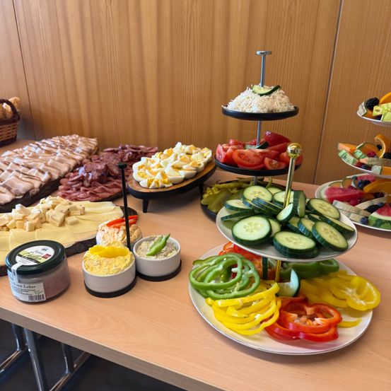 Auf einem Holztisch befinden sich verschiedene Lebensmittel, darunter ein Käseteller, geschnittene Gurken, Tomaten und ein dreistufiger Teller mit verschiedenen Gemüsesorten. Es gibt auch ein Glas Aufstrich und zwei kleine Schüsseln. Im Hintergrund ist eine Holzwand zu sehen.