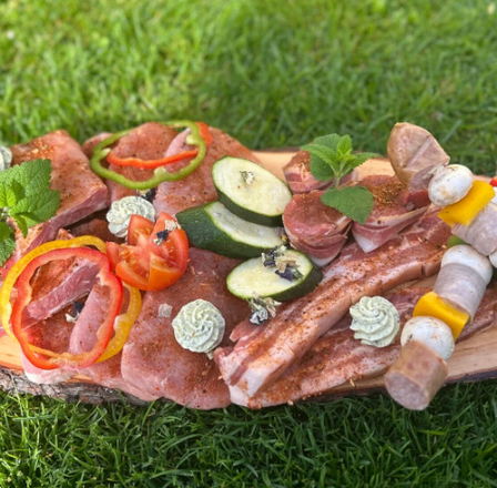 Ein Holzbrett hält eine Vielzahl von Fleisch, Gemüse und Spießen auf einem Rasen. Es enthält geschnittenes Fleisch, Pfefferringe, Gurkenscheiben und Pilzspieße.
