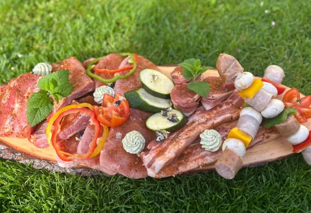 Ein Holzbrett hält eine Vielzahl von Fleisch, Gemüse und Spießen auf einem Rasen. Es enthält geschnittenes Fleisch, Pfefferringe, Gurkenscheiben und Pilzspieße.