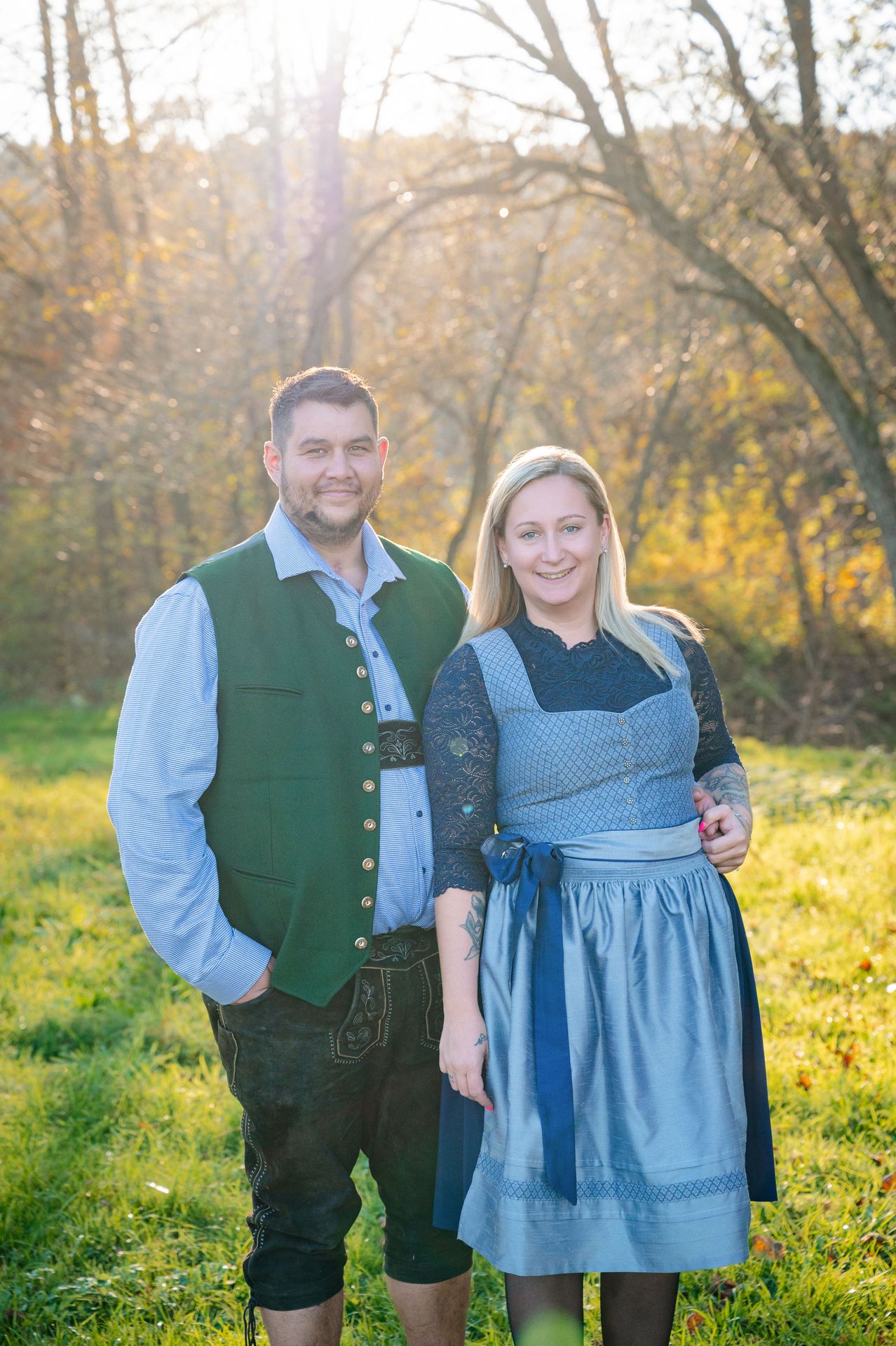 Ein Mann und eine Frau stehen lächelnd zusammen auf einer Wiese. Der Mann trägt eine grüne Weste über einem blauen Hemd, während die Frau ein blaues Kleid mit Spitzendetails trägt. Hinter ihnen stehen Bäume mit Herbstlaub.