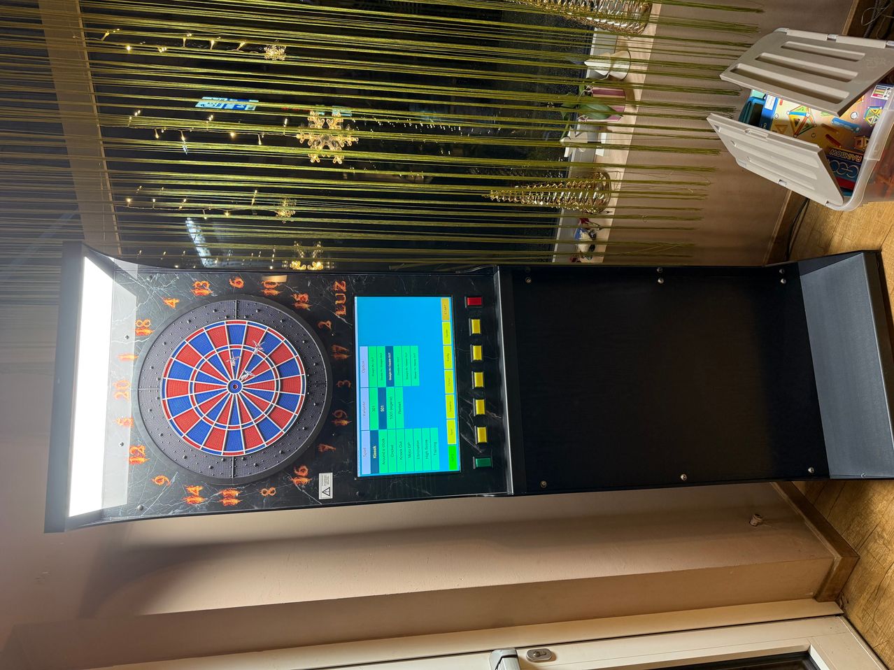Ein schwarzes elektronisches Dartboard mit Digitalanzeige, Zahlen und Tasten ist an einer Wand angebracht.