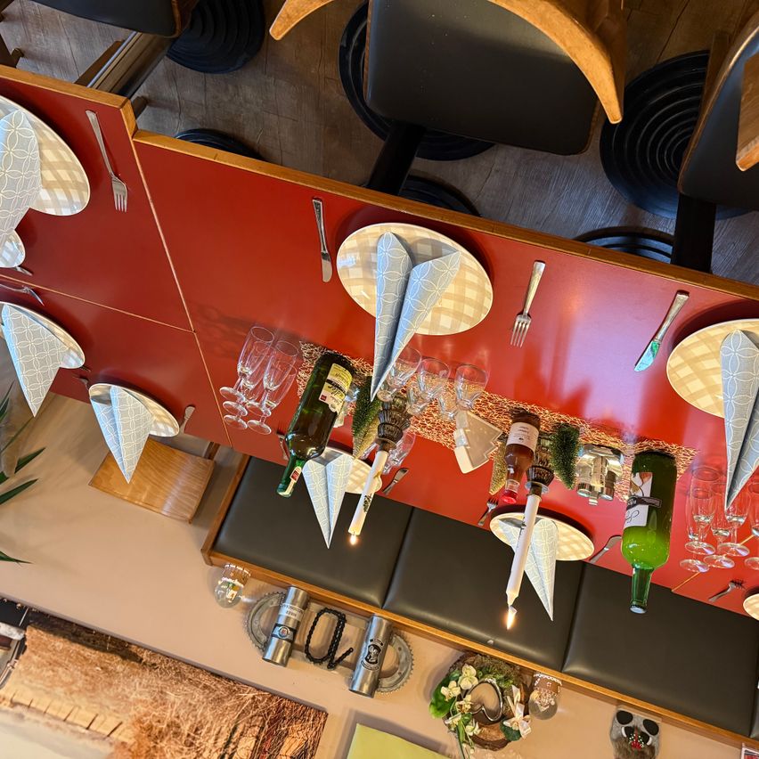 Eine Draufsicht auf einen Tisch, der mit Weingläsern, Flaschen, Tellern, Servietten und Besteck gedeckt ist. Der Tisch hat eine rote Oberfläche und hölzerne Kanten. Die Tischdekoration scheint für eine formelle Veranstaltung vorbereitet zu sein.