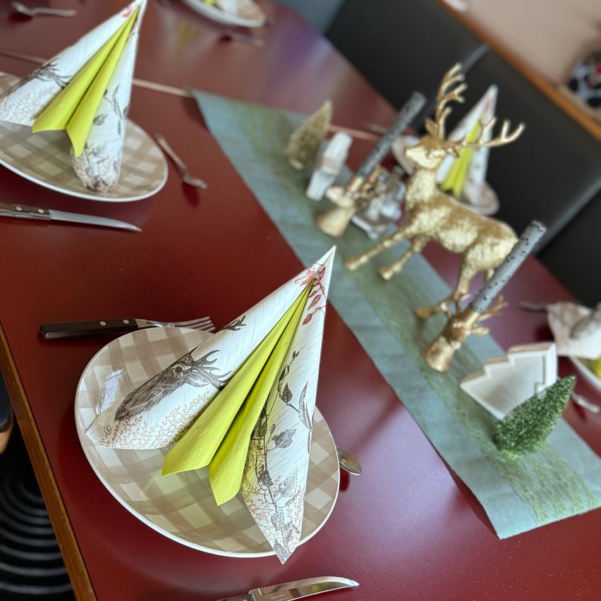 A red dining table is set with festive decorations. Each plate has a folded napkin and a deer figurine. The table has a green runner with Christmas tree decorations. Silverware and knives are placed neatly. The atmosphere is elegant and festive.