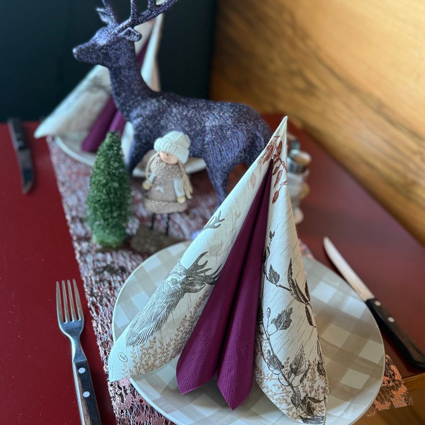 A table setting with folded napkins, a red table runner, a small Christmas tree, a figurine, and a deer statue. A knife, fork, and salt and pepper shakers are also present.