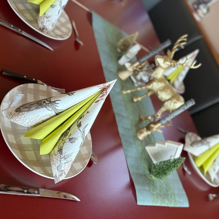 Eine festliche Tischdekoration mit Tellern, zu Dreiecken gefalteten Servietten, Besteck und Rentierfiguren. Der Tisch ist rot mit einem grünen Tischläufer und kleinen Tannenbäumen.