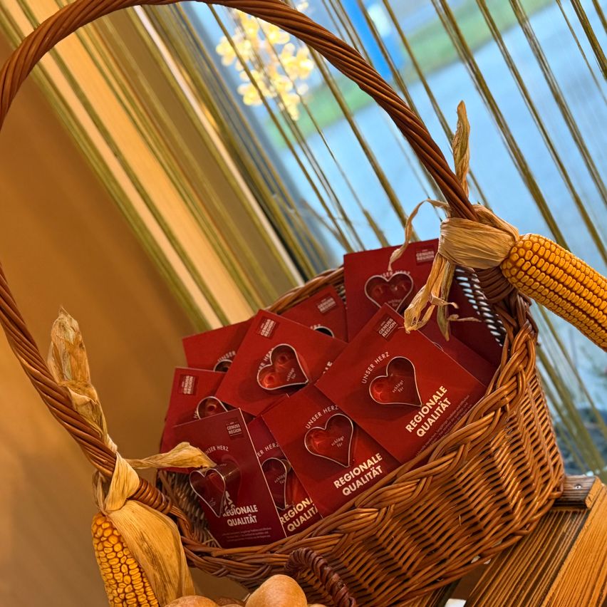 Ein Korb aus Weide gefüllt mit roten herzförmigen Karten, beschriftet mit 'Regionale Qualität', steht neben einer Maiskolbe und einem goldenen Zierlicht.