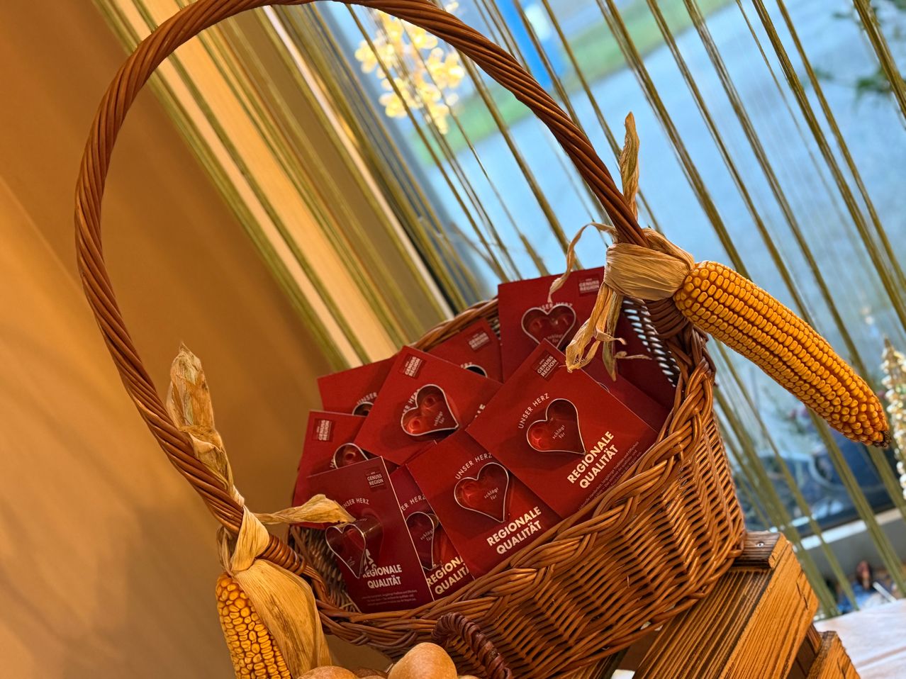 Ein Korb aus Weide gefüllt mit roten Karten mit Herzdesign, auf einer Holzoberfläche mit einem Maiskolben platziert. Die Karten stehen für 'Regionale Qualität' und sind von getrockneten Blättern umgeben.