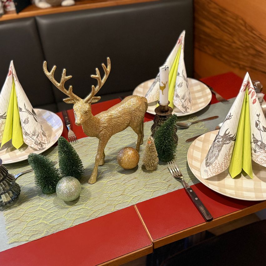 Eine festliche Tischdekoration mit einem goldenen Rentier als Mittelpunkt, Weihnachtsbäumen und dekorativen Tellern. Jeder Teller hat eine grüne Serviette mit Hirschdesign und eine Kerze. Der Tisch hat eine rote Tischdecke.