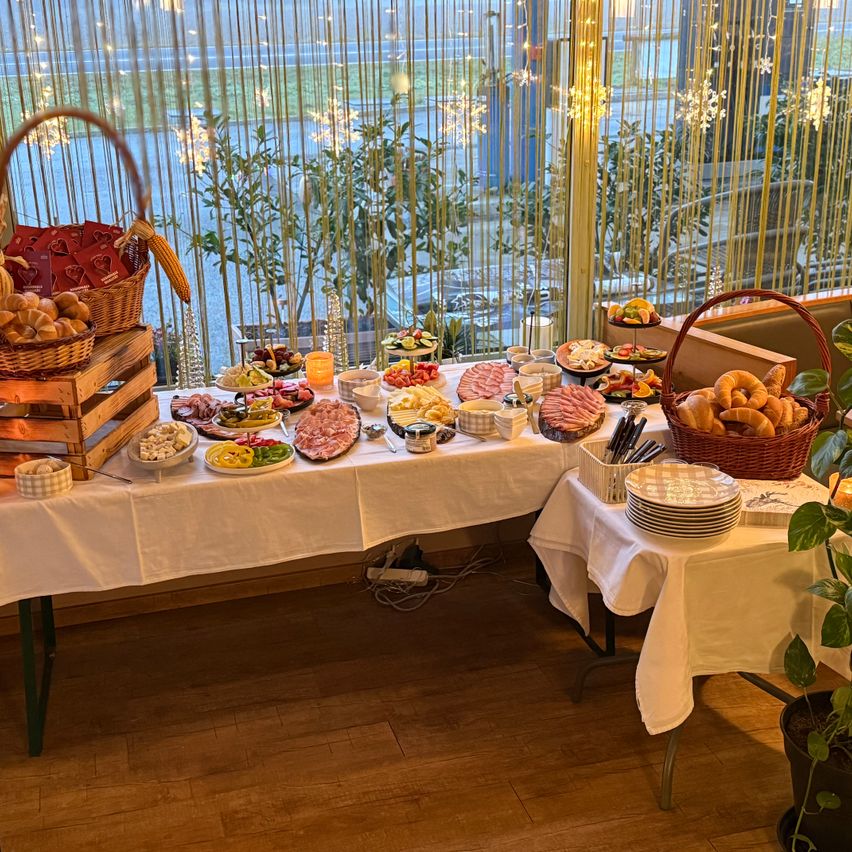 Ein Buffettisch ist mit einer Vielzahl von Lebensmitteln wie Fleisch, Obst und Broten eingerichtet und befindet sich in einem Raum mit Blick auf das Freie und Pflanzen.