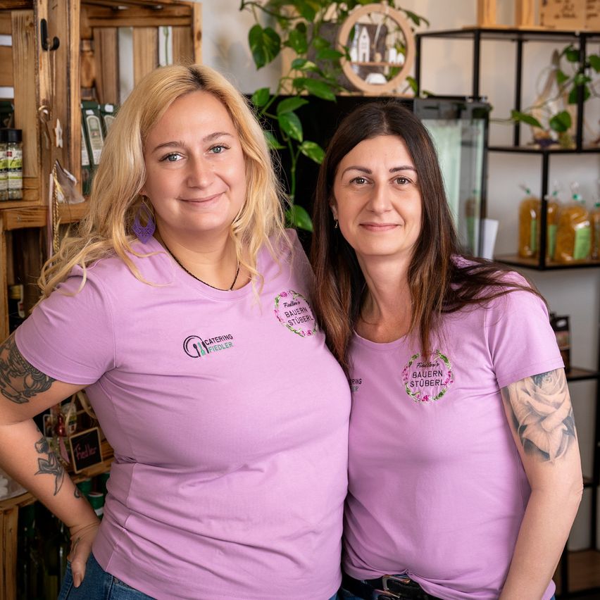 Zwei Frauen in lila T-Shirts mit Blumenlogos posieren für ein Foto in einem Raum mit Holzregalen, Pflanzen und Flaschen.