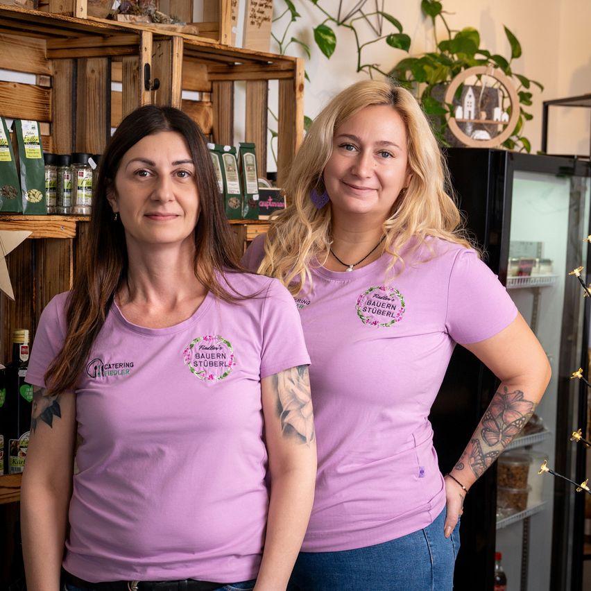 Zwei Frauen stehen nebeneinander und tragen passende lila T-Shirts mit Logos, lächeln vor einem Laden. Dahinter befinden sich Regale mit verschiedenen Produkten und ein schwarzer Kühlschrank ist sichtbar.